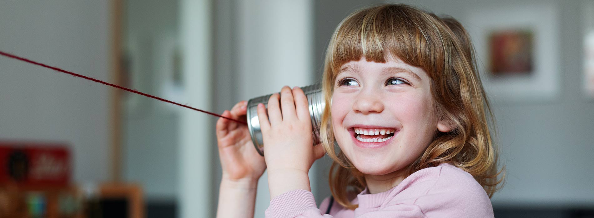 Foto: ein kleines lachendes Mädchen hält eine Dose aus Blech an ihr Ohr und spielt Telefonieren