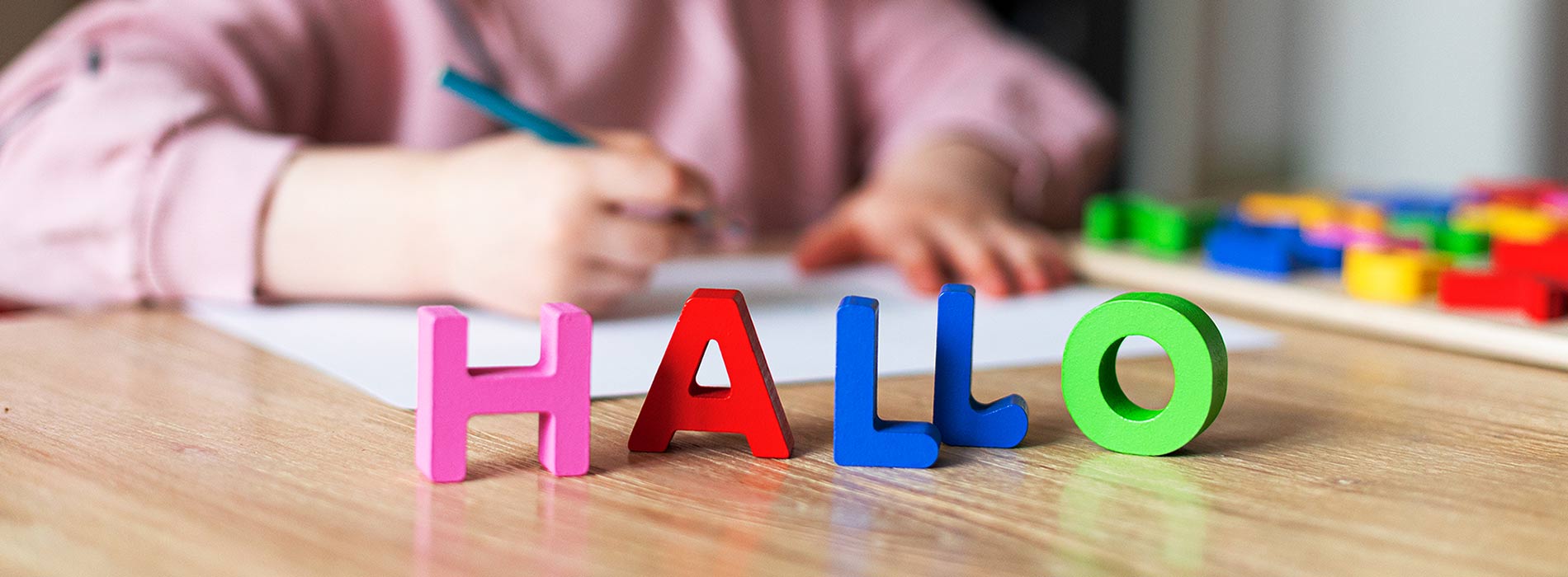 Foto: auf einem Tisch steht das Wort Hallo in bunten Holzbuchstaben und im Hintergrund malt ein kleines Kind mit Buntstift